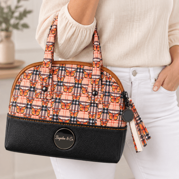 Model wearing an ivory blouse and white jeans carrying a compact orange butterfly plaid handbag with black faux leather base.
