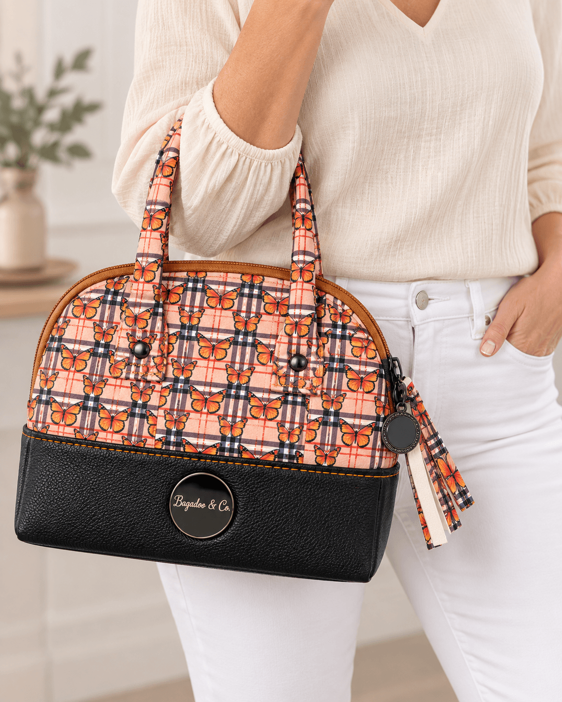 Model wearing an ivory blouse and white jeans carrying a compact orange butterfly plaid handbag with black faux leather base.